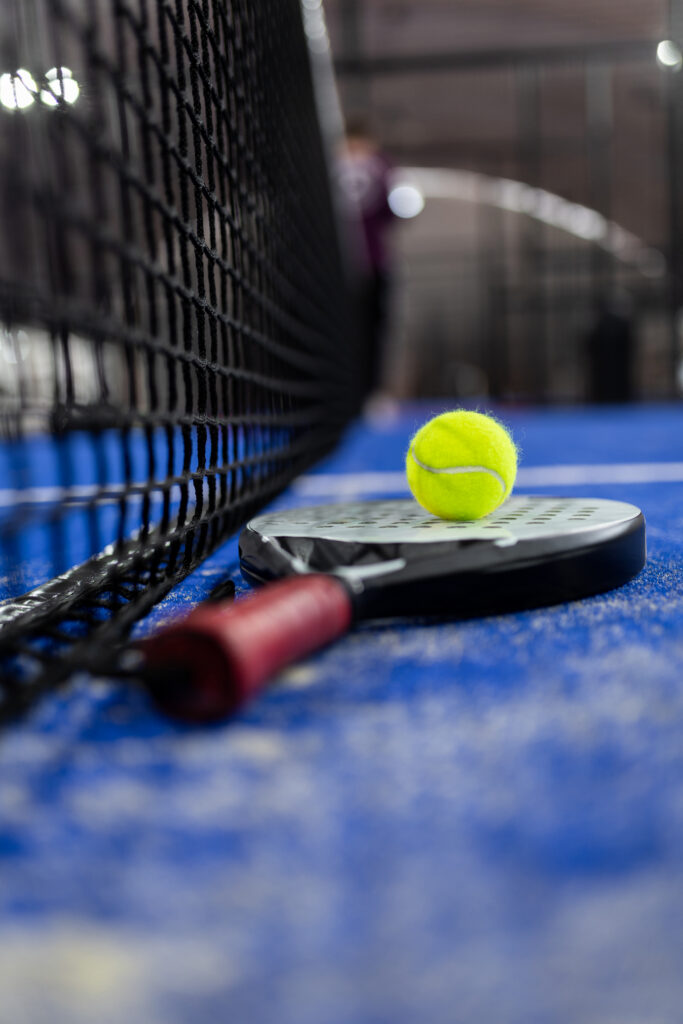 A padel or paddle tennis racket and a bright yellow tennis ball lie on a blue court.